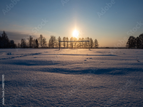 Snow sparkles at snowy football field in winter evening. Silhouette of trees in rays of the setting sun. Place for your text. The cold season.