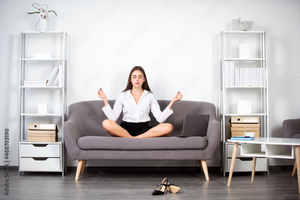 Young meditation businesswoman, secretary girl doing yoga exercise on sofa at workplace in a modern office. Employee feel balance harmony relaxation.