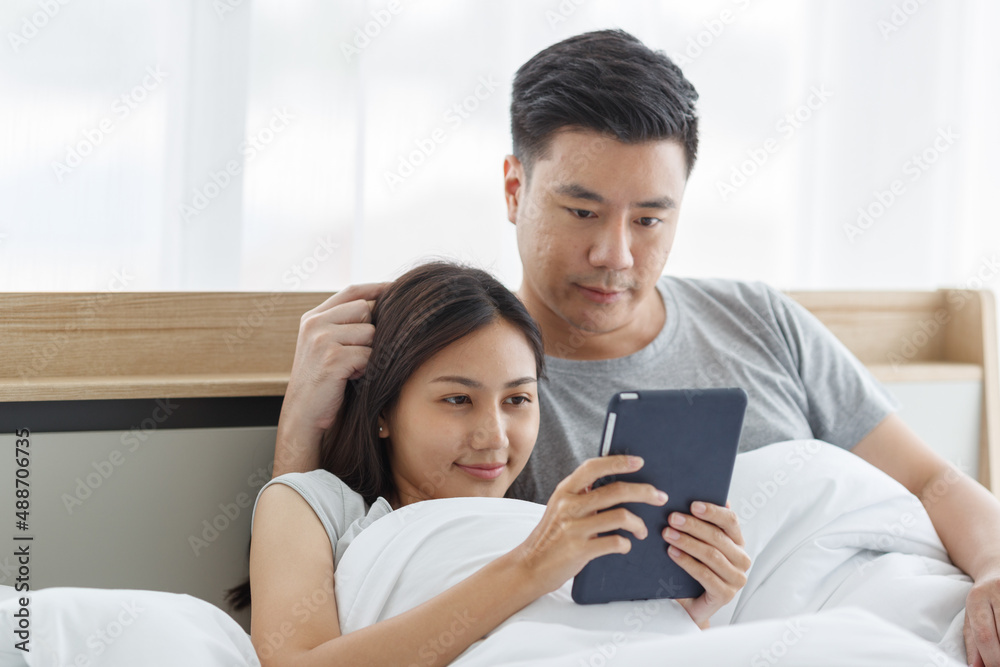 Asian lover couple husband and wife laying down smiling hugging cuddling together on bed in white warm blanket watching online streaming movie via touchscreen tablet computer in bedroom in morning Asian lover couple husband and wife laying down smiling hugging cuddling together on bed in white warm blanket watching online streaming movie via touchscreen tablet computer in bedroom in morning