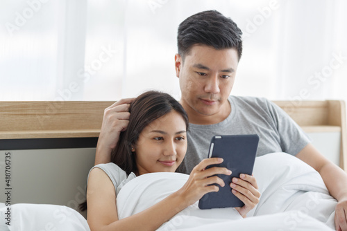 Asian lover couple husband and wife laying down smiling hugging cuddling together on bed in white warm blanket watching online streaming movie via touchscreen tablet computer in bedroom in morning