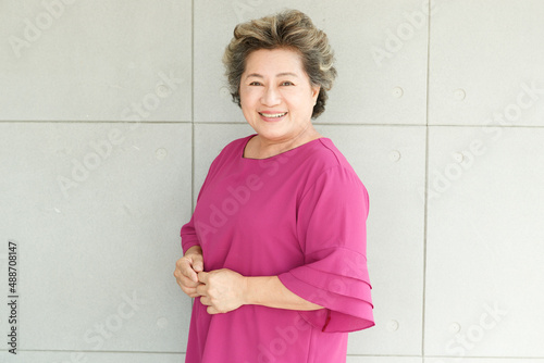 Portrait shot of Asian old senior chubby female retirement pensioner gray short hair grandmother model in casual pink outfit standing smiling look at camera holding hands together on wall background