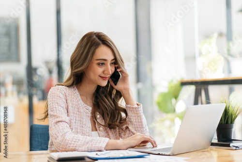 Young happy attractive beautiful asia female staff laugh talk on mobile phone relax comfort sit work on desk table at home. Remotely work on distance job telesale or secretary career for business.