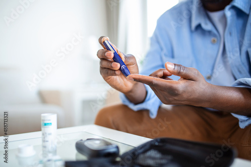 African man is sitting at the sofa at the home and taking blood from his finger due to diabetes. photo