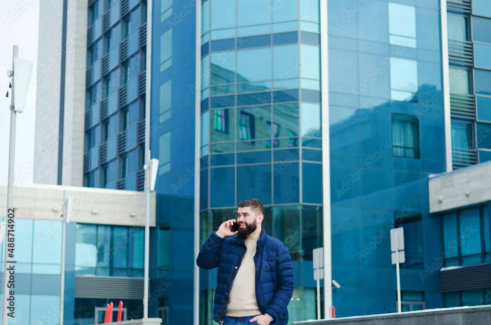Urban portrait of young elegant business man in winter casual clothes, jacket, hat. Walking in the city street, talking on the phone, making selfie. Working outdoor