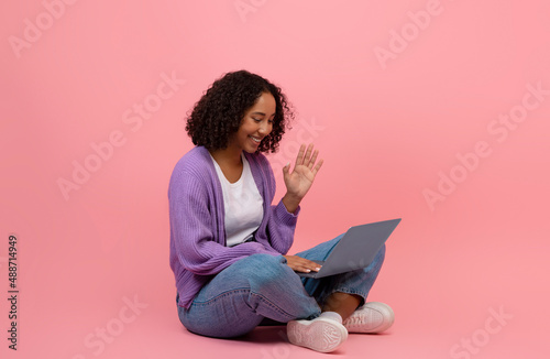 Full length of young black woman sitting cross legged with laptop, communicating online, waving at webcam