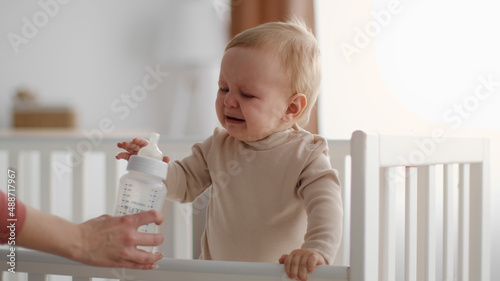 Bild auf Leinwand Cute little unhappy baby crying in crib, mother hand giving bottle with water or