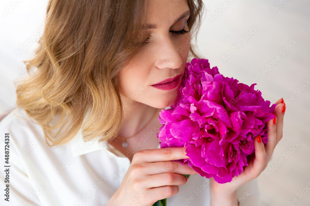 Fototapeta premium woman in white shirt, holding peony bud in her hands, closed her eyes. Portrait of beautiful blonde woman. Surprise bouquet of flowers for date, mothers day or valentines day.