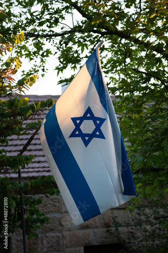 A large, blue and white flag of the State of Israel hanging from a pole outside a home on a sunny day in Israel.