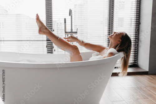 Playful young lady shaving legs with razor, lying in foamy bubble bath, enjoying domestic skin care procedure, side view photo