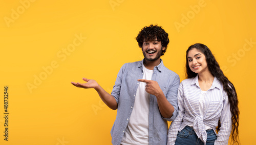 Cheerful indian couple pointing at open palm, man demonstrating invisible object, showing copy space, panorama photo