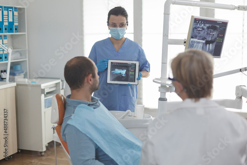 Medical nurse holding tablet computer explaining radiography diagnosis discussing teeth treatment with patient during stomatology consultation in dental office. Dentistry team wearing face mask