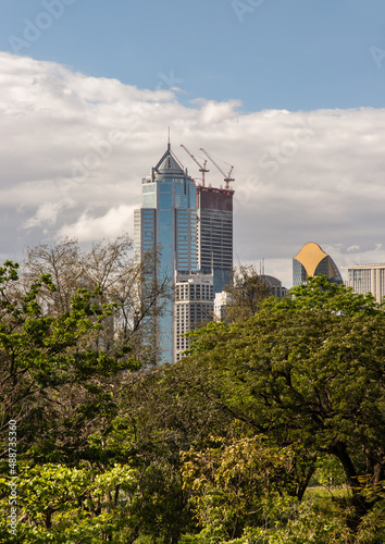 View of Modern high buildings among green trees space in nature against blue sky with clouds at afternoon. City growth concept, Selective focus.