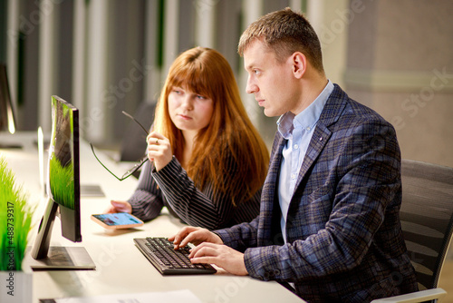 Coworking, a young woman and a business man are discussing a project looking at a laptop monitor and screen, studying documentation, discussing project activities, business partners.