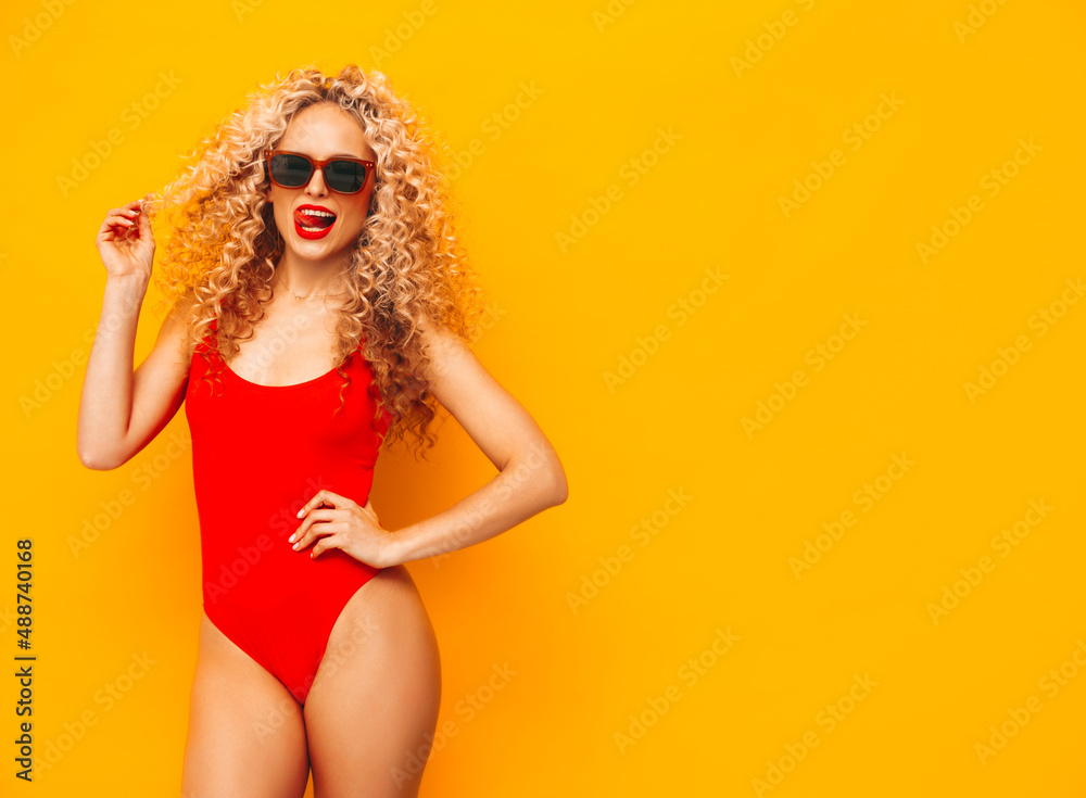 Young beautiful smiling woman posing near yellow wall in studio.Sexy model in red swimwear bathing suit.Positive female with curls hairstyle.Happy and cheerful. In sunglasses Young beautiful smiling woman posing near yellow wall in studio.Sexy model in red swimwear bathing suit.Positive female with curls hairstyle.Happy and cheerful. In sunglasses
