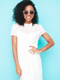 Portrait of beautiful black woman with afro curls hairstyle. Smiling model dressed in white summer dress. Sexy carefree female posing near blue wall in studio. Tanned and cheerful