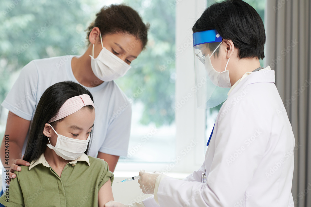 Fototapeta premium Doctor in protective wear giving a vaccination to little girl while she visiting the hospital together with her sister