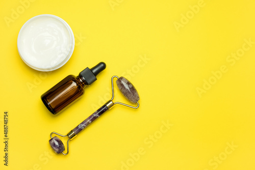 Face roller for beauty facial massage therapy, Items for home treatment with Skin Care Anti-Aging Tools, Face Serum and Moisturiser Cream on yellow background, top view. © elenae333
