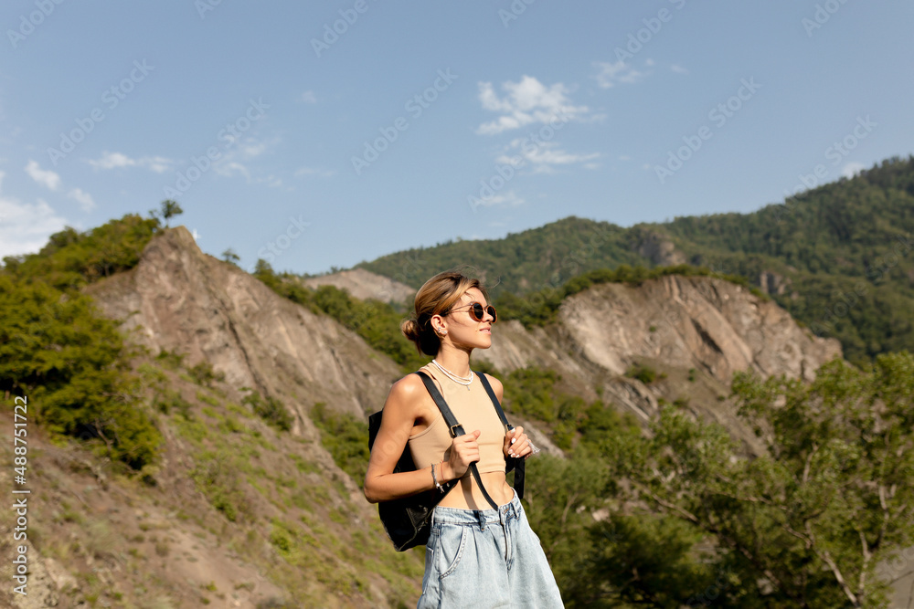 Adorable charming lady with wavy light hair wearing t-shirt and shorts with backpack is moving against green mountains in sunlight in warm spring day. Woman traveling in Woman's Day Adorable charming lady with wavy light hair wearing t-shirt and shorts with backpack is moving against green mountains in sunlight in warm spring day. Woman traveling in Woman's Day