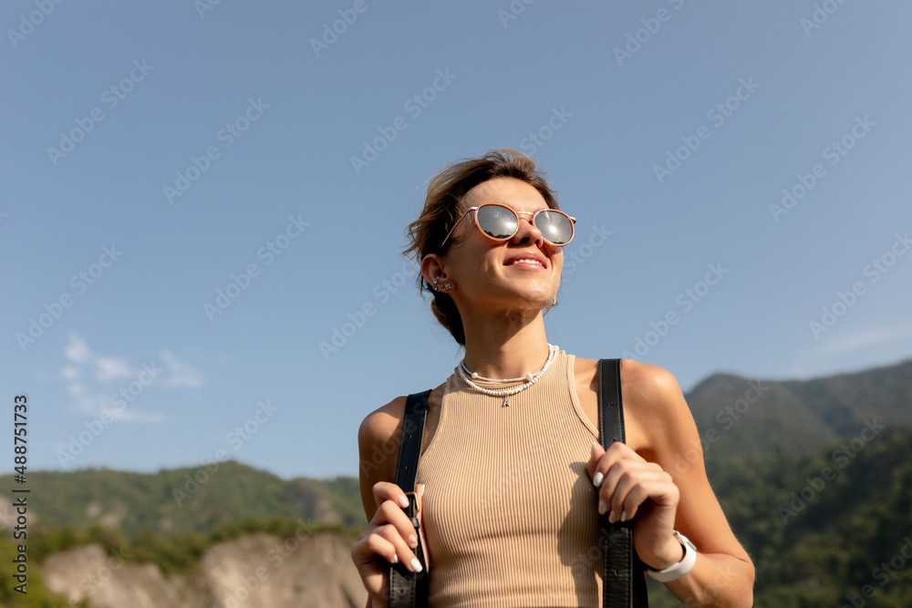Close up portrait of attractive charming woman with collected light hair wearing sunglasses and beige t-shirt is walking aside in mountains on background of blue sky