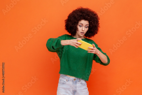 Young adult woman with Afro hairstyle wearing green casual style sweater playing online video game with scared and concentrated facial expression. Indoor studio shot isolated on orange background.