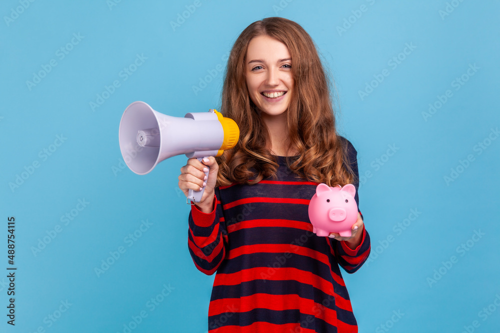 Portrait of positive woman wearing striped casual style sweater, holding piggy bank and megaphone, looking at camera with toothy smile. Indoor studio shot isolated on blue background. Portrait of positive woman wearing striped casual style sweater, holding piggy bank and megaphone, looking at camera with toothy smile. Indoor studio shot isolated on blue background.