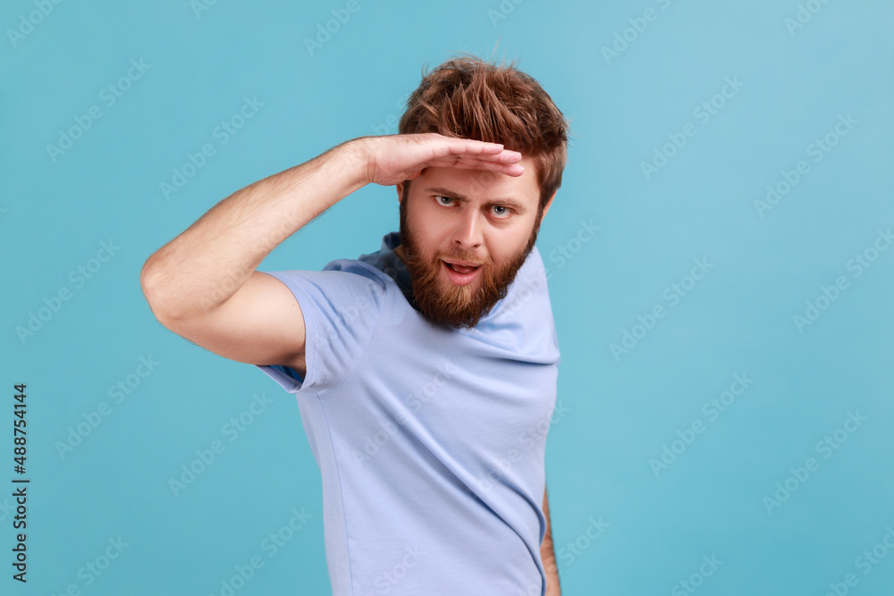Portrait of handsome young adult bearded man looking far away at distance with hand over head, sees something pleasant with flirting expression. Indoor studio shot isolated on blue background. Portrait of handsome young adult bearded man looking far away at distance with hand over head, sees something pleasant with flirting expression. Indoor studio shot isolated on blue background.
