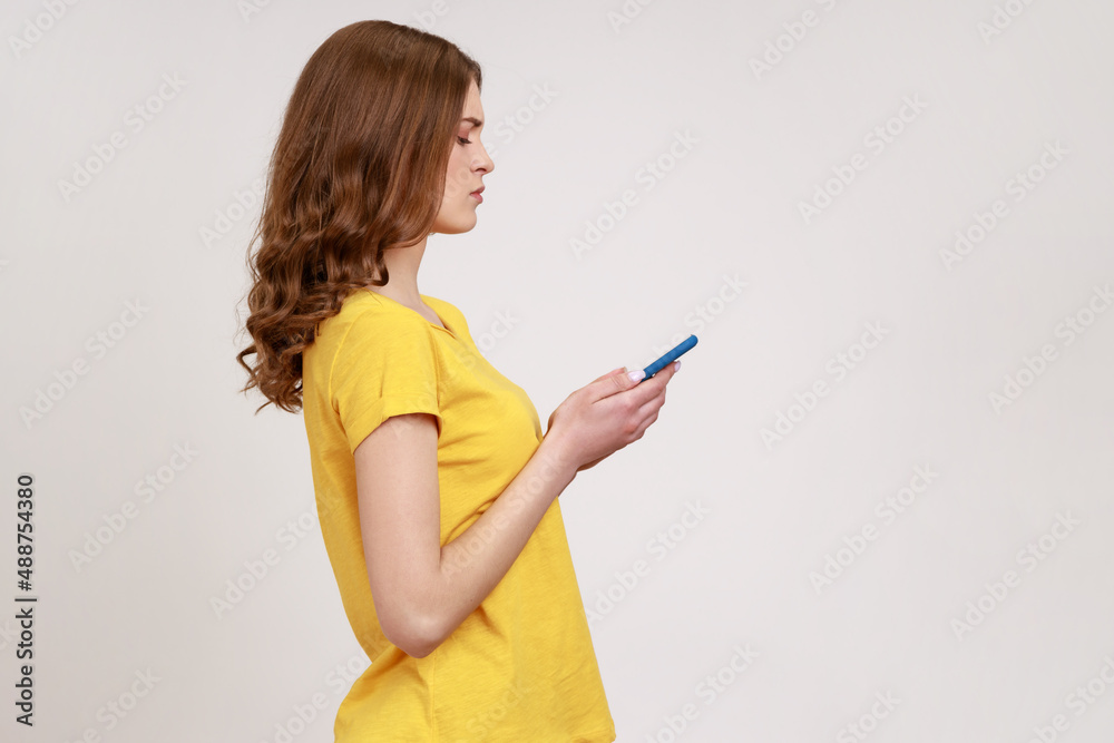 Side view of serious attentive teen girl typing on cell phone, reading blog, chatting in social network with friends, using mobile app for communication. Indoor studio shot isolated on gray background Side view of serious attentive teen girl typing on cell phone, reading blog, chatting in social network with friends, using mobile app for communication. Indoor studio shot isolated on gray background