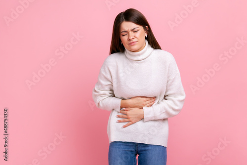 Ill brunette woman suffering acute pain, cramps from indigestion or gastritis, appendicitis symptoms, wearing white casual style sweater. Indoor studio shot isolated on pink background.
