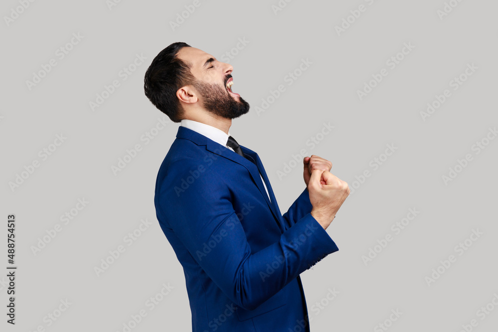 Side view of bearded man showing yes gesture and screaming celebrating his victory, success, dreams comes true, euphoria, wearing official style suit. Indoor studio shot isolated on gray background. Side view of bearded man showing yes gesture and screaming celebrating his victory, success, dreams comes true, euphoria, wearing official style suit. Indoor studio shot isolated on gray background.