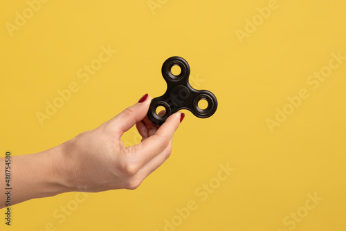 Closeup side view profile portrait of woman hand holding black fidget spinner, stress relieving toy. Indoor studio shot isolated on yellow background. photo