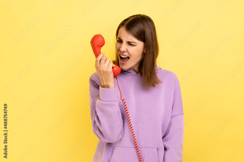 Housing maintenance call center. Angry brown haired woman shouting into retro phone handset, irritated by customer call, wearing purple hoodie. Indoor studio shot isolated on yellow background. Housing maintenance call center. Angry brown haired woman shouting into retro phone handset, irritated by customer call, wearing purple hoodie. Indoor studio shot isolated on yellow background.
