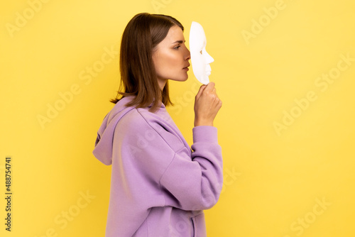Side view of anonymous dark haired woman covering her face with white mask, hiding personality, conspiracy and privacy, wearing purple hoodie. Indoor studio shot isolated on yellow background.