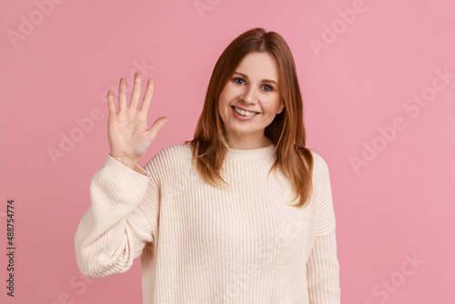 Portrait of young blond friendly woman waving hand saying hi welcome, smiling with hospitable sociable expression, wearing white sweater. Indoor studio shot isolated on pink background.