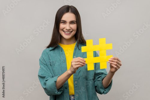 Portrait of happy beautiful woman holding hashtag symbol, promoting viral topic in social network, tagging blog trends, wearing casual style jacket. Indoor studio shot isolated on gray background.