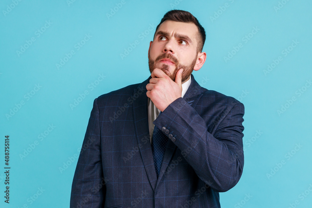 Confused uncertain man with beard wearing dark official style suit holding his chin choosing, trying to make right decision, dilemma. Indoor studio shot isolated on blue background.