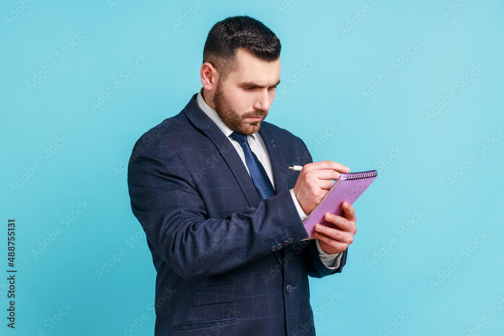 Bearded handsome man wearing dark official style suit writing down thoughts holding diary, making notes at notepad, writing to do list. Indoor studio shot isolated on blue background. Bearded handsome man wearing dark official style suit writing down thoughts holding diary, making notes at notepad, writing to do list. Indoor studio shot isolated on blue background.