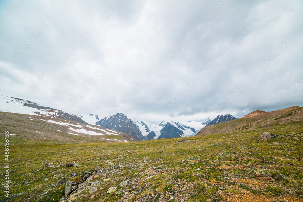 Scenic panoramic view from sunlit green grassy hill to high snowy mountain range with sharp tops and glaciers under gray cloudy sky. Colorful landscape with large snow mountains at changeable weather.