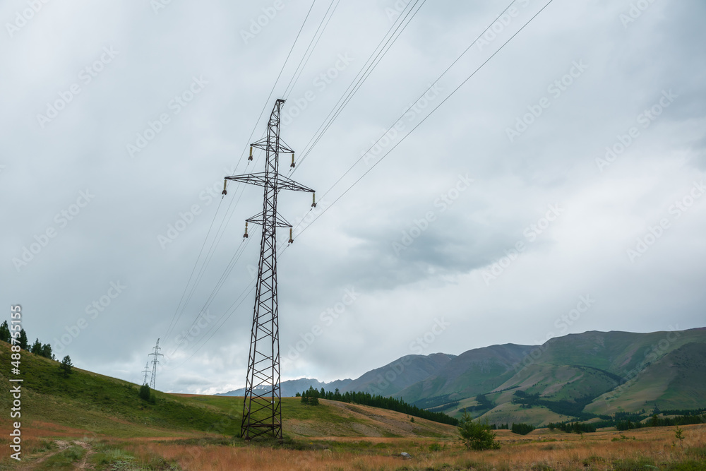 Dramatic mountain landscape with tall tower of power lines under cloudy sky. Atmospheric alpine scenery with electric tower with wires against high mountains in overcast. Electricity in mountains.