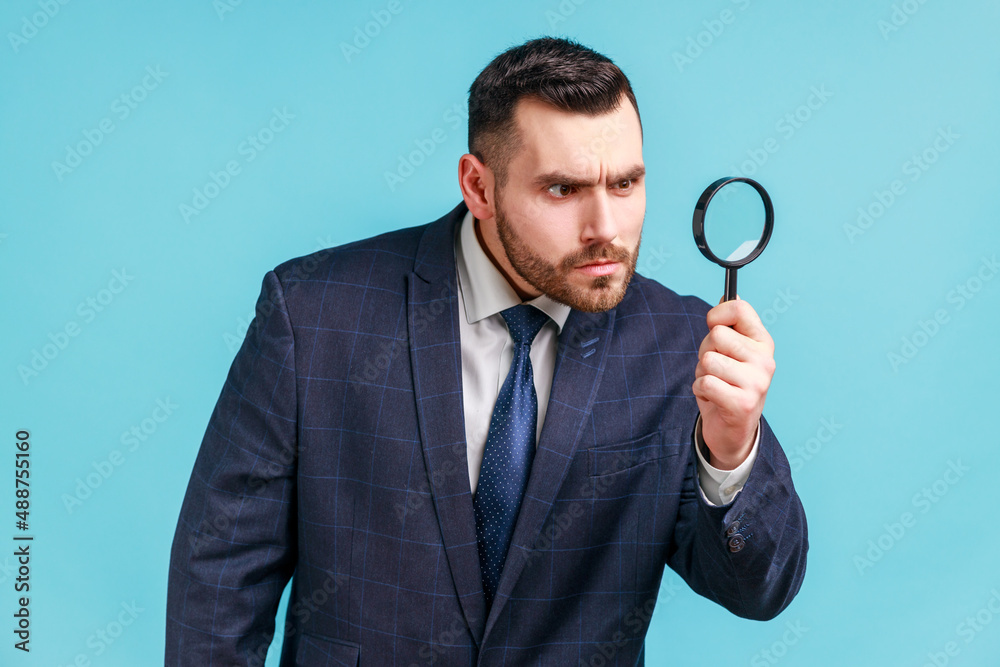 Serious man with beard wearing in dark suit looking away through magnifying glass, spying, finding out something, exploring crime scene, inspecting. Indoor studio shot isolated on blue background. Serious man with beard wearing in dark suit looking away through magnifying glass, spying, finding out something, exploring crime scene, inspecting. Indoor studio shot isolated on blue background.