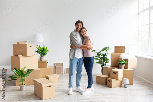 Glad young caucasian male and female in casual hugging in room with cardboard boxes, renting flat and relocation