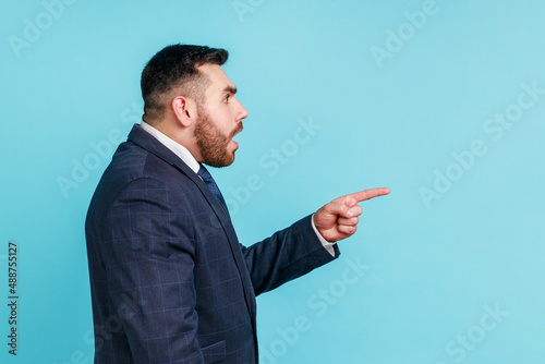 Portrait of attentive handsome bearded young man wearing official style suit having shocked facial expression, pointing finger aside. Indoor studio shot isolated on blue background.