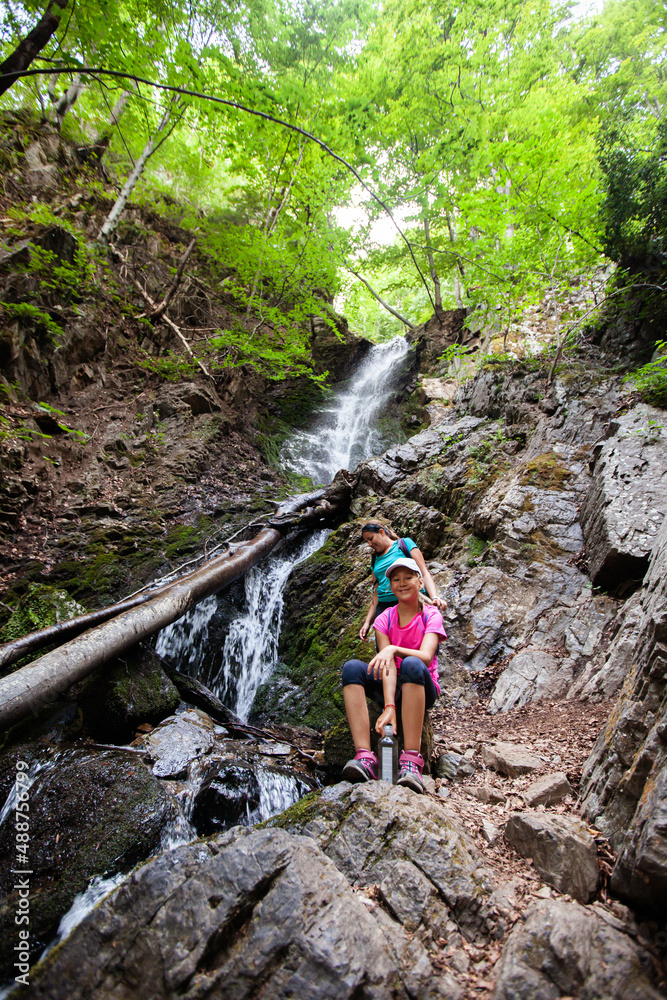Daughter sitting on rock and drink water. Happy mother in background. Mountain waterfall in forest. Nature landscape. Summer season. Day light.