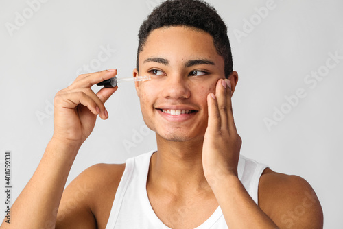 Young African-American guy with serum for skin care on grey background
