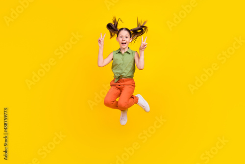 Photo of cute charming preteen girl dressed green top showing two v-signs jumping high smiling isolated yellow color background