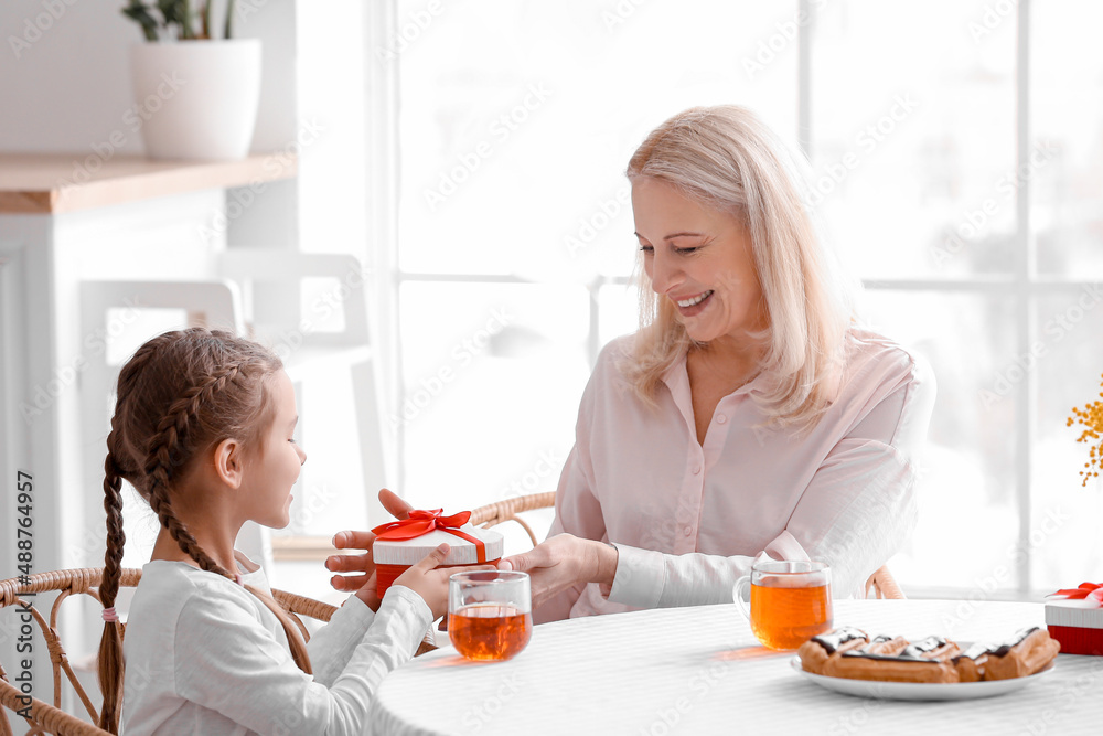 Little girl greeting her grandmother with gift in kitchen on International Women's Day