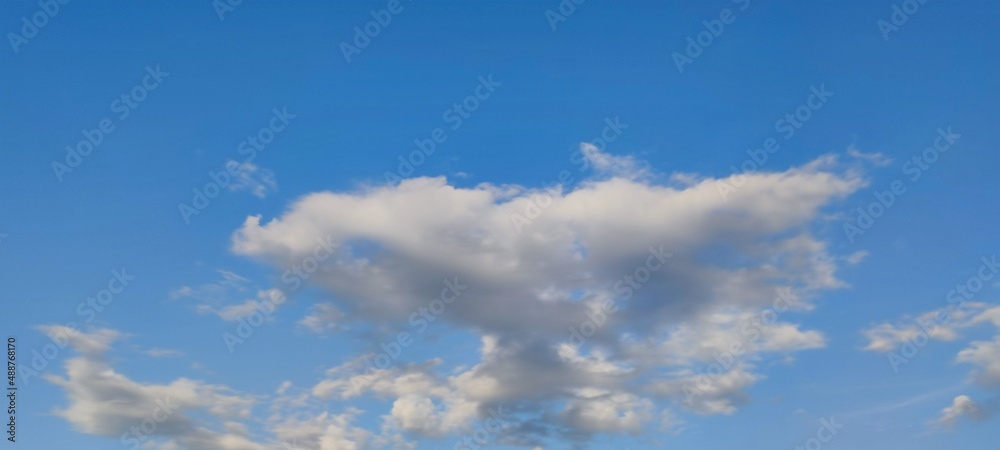 White fluffy cumulus clouds in the sky. Against the background of a dark blue sky, white and slightly gray clouds of various shapes and sizes are floating. The sun illuminates the clouds from one side