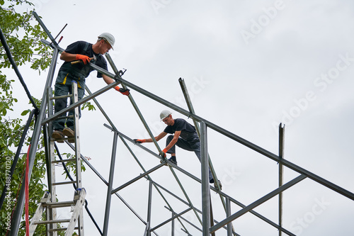 Man in safety helmet standing on ladder and holding hammer while colleague installing metal rail for solar modules. Male workers assembling support structures for solar panels under white sky.