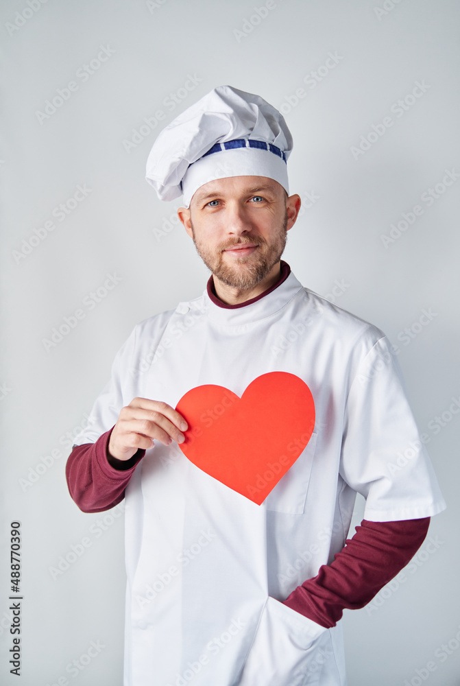 International Chefs Day concept: cheerful bearded male baker or chef in white uniform with red paper heart. Restaurant of cafe concept. Chef cook holding a red heart in front of the workwear International Chefs Day concept: cheerful bearded male baker or chef in white uniform with red paper heart. Restaurant of cafe concept. Chef cook holding a red heart in front of the workwear