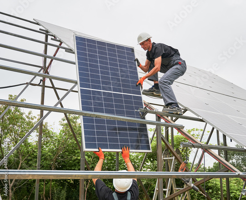 Male worker in safety construction helmet giving solar module to colleague. Two men in workwear building photovoltaic solar panel system. Concept of sustainable energy and solar panel installation.
