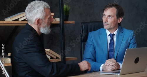 Two mature businessmen discussing business growth in modern office while sitting at desk. Man ceo talking with colleague about deal while meeting indoors